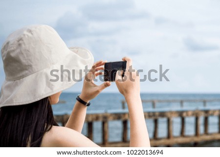 Back view of young woman wearing a hat taking photo with her smart phone on landscape view