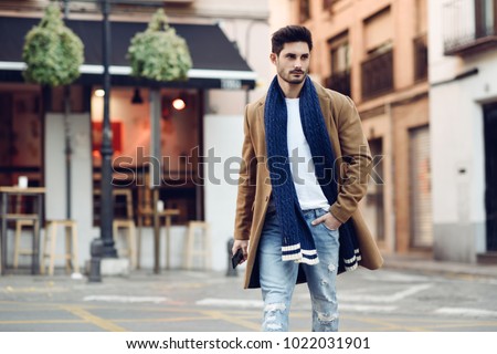 Young man wearing winter clothes with a smartphone in his hand, walking in the street. Young bearded guy with modern hairstyle in urban background.