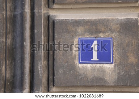 Close up view of the number one written on a small blue rectangle board fixed on the wall of an old house. Decorative lines on the grey stone wall. Numeric white symbol in a city street in France. 