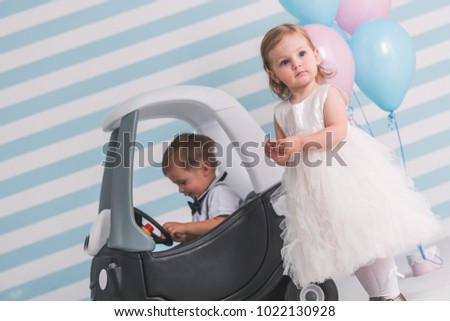 Cute kids in elegant clothes on light background. Boy is sitting in the toy car, girl is standing near