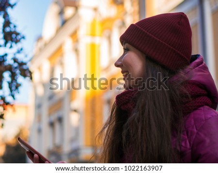 Pretty Brunette Girl Wearing Purple Winter Coat, Hat and Scarf, Walking by European Street at Winter, Using Her Smartphone and Making Selfie