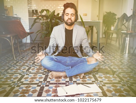 Young businessman in headphones meditating in lotus pose taking a deep breath at living room.Business yoga and stress free environment.Peace of mind concept.Blurred background