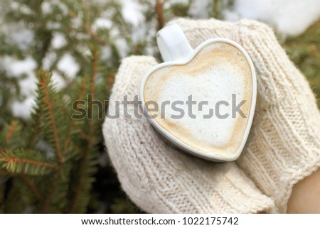 foamy hot cappuccino in the shape of a heart in the hands dressed in knitted mittens against the background of the winter landscape / warming power of love