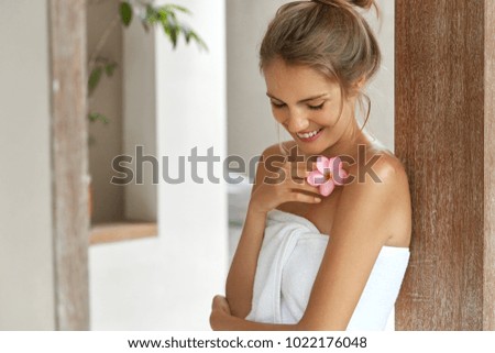 Beautiful young woman with soft skin, has light hair tied in knot, wears white towel after taking douche, smells flower, looks down with happy expression, pleased after good relax at bathroom Royalty-Free Stock Photo #1022176048