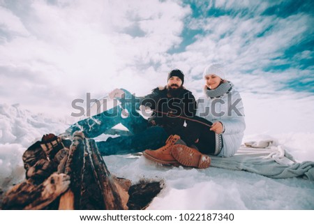 wife in white winter jacket, mitten and hat sitting near to her husband and looks at him. The concept of the happy couple, winter camping, hiking, recreation, tourism Royalty-Free Stock Photo #1022187340
