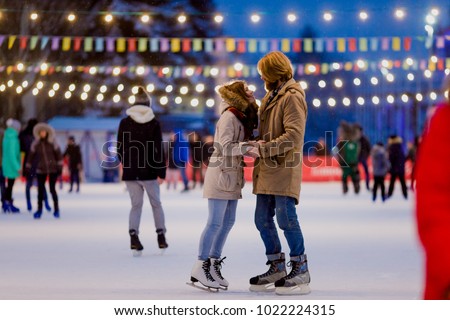 Ice skating rink and lovers together. A pair of young, stylish people in an embrace in a crowd on a city skating rink lit by light bulbs and bright lights. Winter date for Christmas on the ice arena