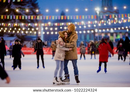 Ice skating rink and lovers together. A pair of young, stylish people in an embrace in a crowd on a city skating rink lit by light bulbs and bright lights. Winter date for Christmas on the ice arena