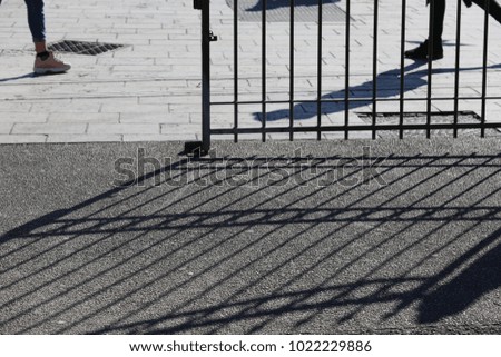 Abstract close up view of the base of a public garden gate and its shadow. Silhouettes with lines and people walking in front of the entrance. Lighted paved surface. Some legs and feet are visible. 