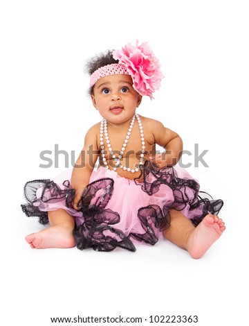 A beautiful six month old baby girl wearing a pink headband with a big flower and a tutu and sitting against a white background