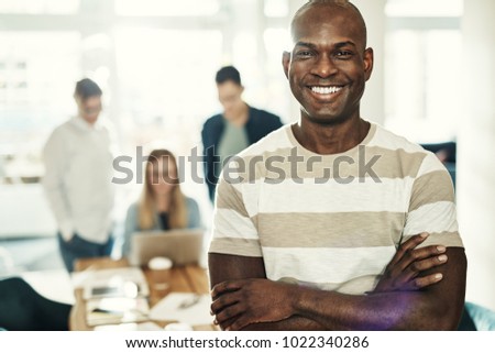 Confident young African designer smiling while standing with his arms crossed in a modern office with colleagues working in the background