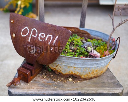 Rustic country Open sign, written in chalk on a rusty vice grip clamp plate. Sitting on an old sodden chair, and supported by a rusted out, holed planter bucket.