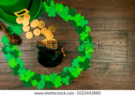 Saint Patrick's Day. Small bag of leprechaun with gold coins lying on wooden background in circle shape of green three petal clovers, gift's box, gold coins, green hat of leprechaun and smoking pipe