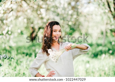 Pretty young happy brunette girl in white shirt with emotional cheerful face smiling, laughing like child in spring blooming garden with bouquet of dandelions. Cute female model posing at nature