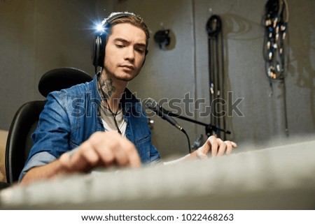 Thoughtful concentrated hipster sound engineer with tattoo on neck listening to song and changing sounds while producing new track in recording studio
