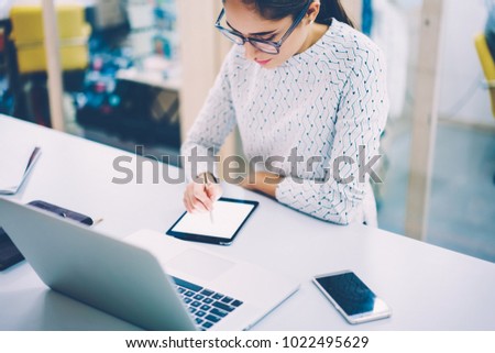 Concentrated female web designer drawing using modern digital gadget for synchronizing sketch with laptop computer,creative woman working as illustrator painting on blank tablet with pen  in office