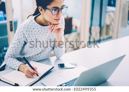 Pensive female administrative assistant checking report on laptop reading information and noting data,skilled woman watching online video online pondering on creating publication writing in notepad Royalty-Free Stock Photo #1022499355