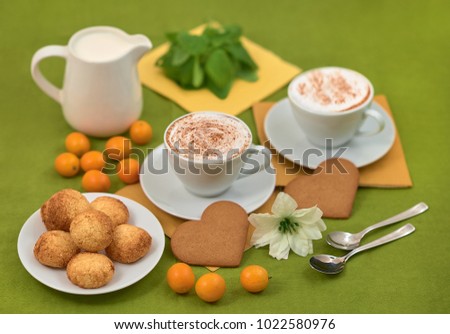 A plate with cookies, two white cups with cappuccino, a milk jug, orange kumquats and a white flower on a green napkin
