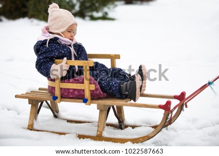 Toddler girl sitting on the sled in the snow, wearing wool gloves and hat and a blue winter suit