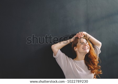 Laughing carefree young redhead woman with her hands raised to her head tilting her face into the sun over a dark grey studio background with copy space