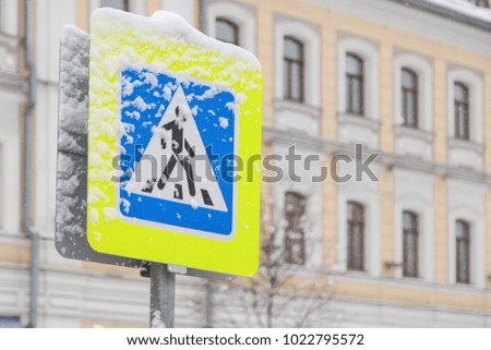 Road sign of a pedestrian crossing covered with snow on a background of houses in the city