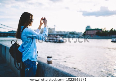 Casually dressed female traveler making pictures of river using smartphone while strolling on bridge in city , young hipster girl shooting video on mobile phone enjoying leisure during warm evening