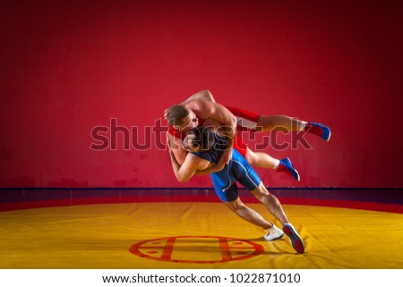 Two young men in blue and red wrestling tights are wrestlng and making a suplex wrestling on a yellow wrestling carpet in the gym