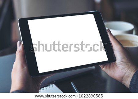 Mockup image of hands holding black tablet pc with blank white desktop screen with coffee cups on table in modern cafe