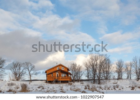 wooden lodge around winter trees and snow, with wooden terrace around home, blue sky, frozen lake.