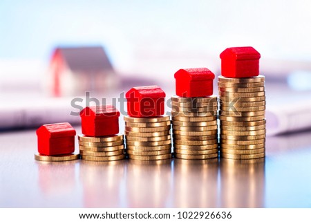 Small red houses standing on stacks of coins with blueprints and architectural model in the background. Royalty-Free Stock Photo #1022926636