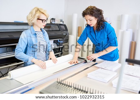 Portrait of two modern young women working in printing shop or publishing company, loading machines with paper and cutting brochures Royalty-Free Stock Photo #1022932315