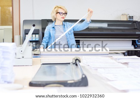 Portrait of blonde young woman working in modern printing shop or publishing company, loading machines with paper, copy space