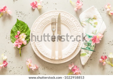 Easter, spring or summer table setting design captured from above (top view, flat lay). Ornamental white plates, glass, cutlery and pink flowers. Grey stone background.