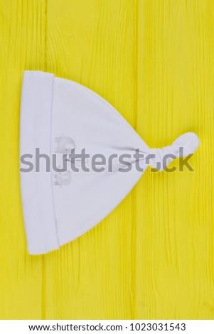 Horizontal image of knotted hat. White cute bayb hat lying on yellow wooden table.