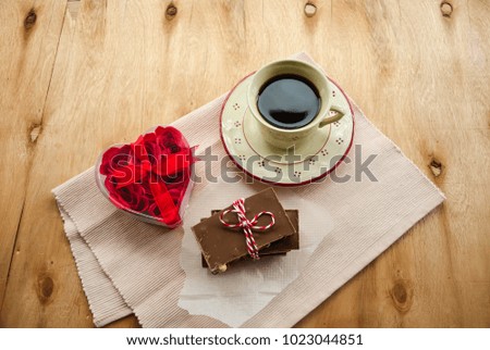A cup of coffee and a white rose with a red heart shape and pieces of chocolate on rustic wooden table 