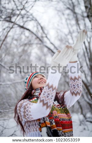 Photo of smiling brunette looking up in knitted hat and scarf catching snowflakes in winter forest during day