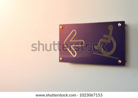 Man in wheelchair sign with arrow pointing forward on navy label,Disabled way symbol on white cement wall and soft flare,copy space for text.Concept is A path for those who have hope.