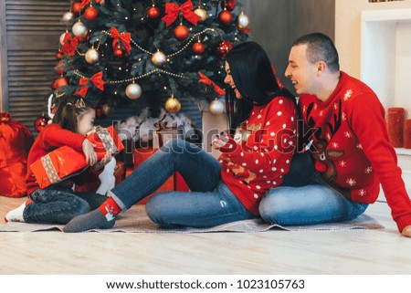 Happy young family of dad, mom and their little daughter dressed in the same red sweaters poses before a Christmas tree in the room