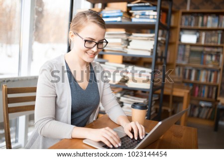 young smiling caucasian woman in glasses  sitting at a table with a laptop on the background of a window and bookshelves