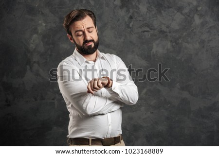 Image of concentrated bearded businessman standing over grey wall background. Looking at watch.