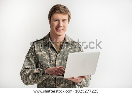 Glad military young man looking at camera with calm smile. He is holding laptop in hand and typing. Isolated on background