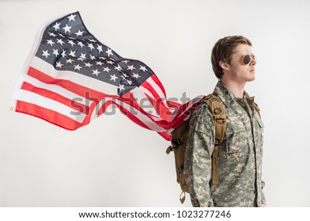 Severe military young man walking somewhere with serious look. He is having backpack on his shoulders and usa flag is fluttering behind his back. Isolated on background