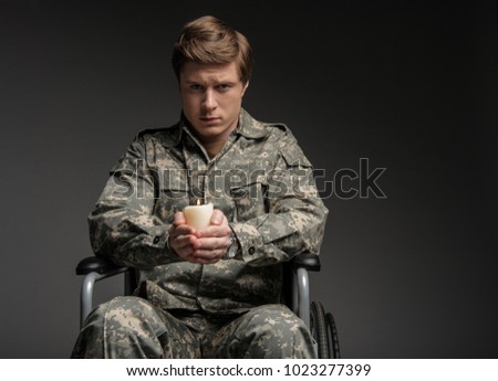 Unhappy paralyzed soldier looking at camera with frustrated look. He is sitting in invalid chair and holding suppository. Isolated on grey background. Copy space in right side