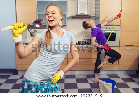 Modern kitchen - woman pretend to sing song with ladle and smiling young man cleaning the floor at home and play like guitar with  mop Royalty-Free Stock Photo #102331519
