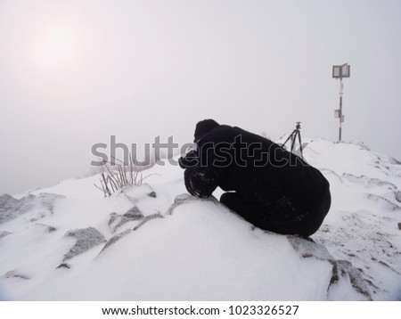 Professional photographer lay down to snow and takes photos with mirror camera on peak of snowy rock. . Man lay with big mirror camera on neck. Snowy rocky peak of mountain.