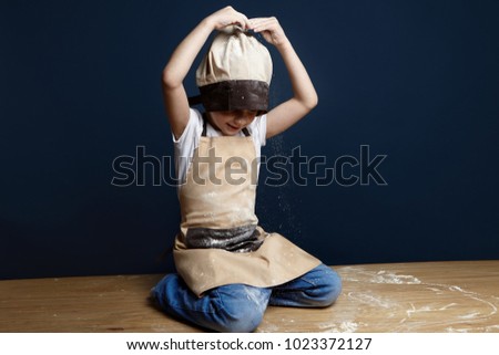 Picture of naughty little boy of Caucasian appearance playing at home sitting on floor in chef headwear, jeans and apron and pouring wheat flour over his head while helping mother to bake cake
