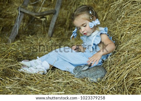Little pretty girl is sitting in the hay with a little rabbit. Girl is feeding her favourite pet. Summer in a village. Happy childhood