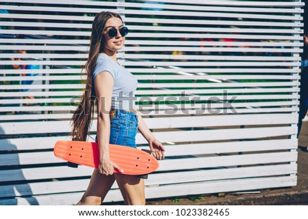Cheerful young woman with long hair and black sunglasses smiling at camera while holding orrange skateboard in hands.Positive sportive young woman strolling near publicity area for advertising