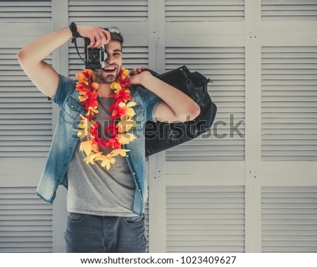 Handsome young man is preparing for traveling. Standing on grey background with suitcase in Hawaii accessories and sunglasses with retro camera in hand.