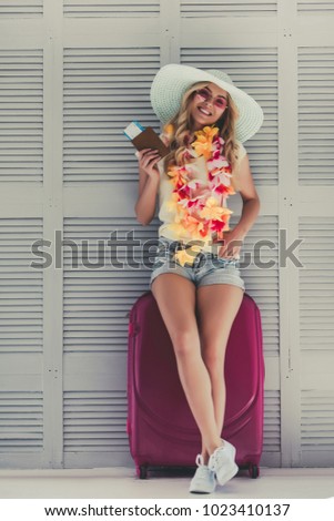 Attractive young woman is preparing for traveling. Standing on grey background with big suitcase in Hawaii accessories, pink sunglasses and hat with passport and ticket in hand.