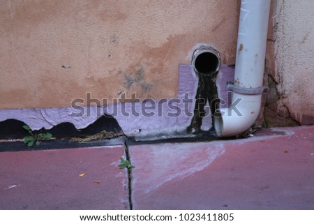 Close up exterior view of the base of a metallic gutter fixed to an orange painted wall. Purple pavement. Dirty water visible. Colorful picture with different pastel tones. Abstract urban image. 